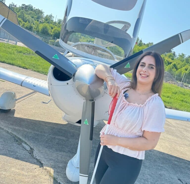 Parimah Attarian står framför propellern på nosen av ett mindre vitt flygplan ute på en landningsbana. Hon har långt mörkbrunt hår, ljusrosa top, bruna tights och håller i en vit käpp.